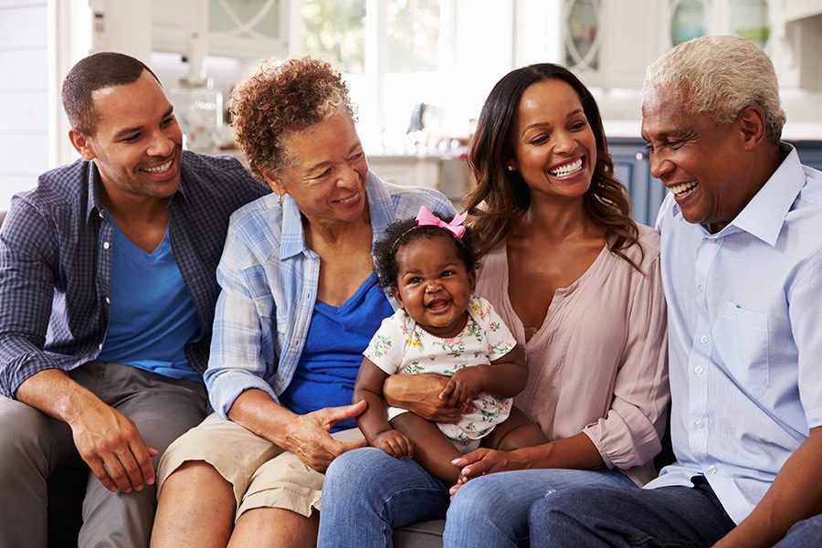 About Our Agency - Smiling Grandparents and Parents Sitting Together on a Sofa With a Baby Girl on Sitting on the Mother’s Knee